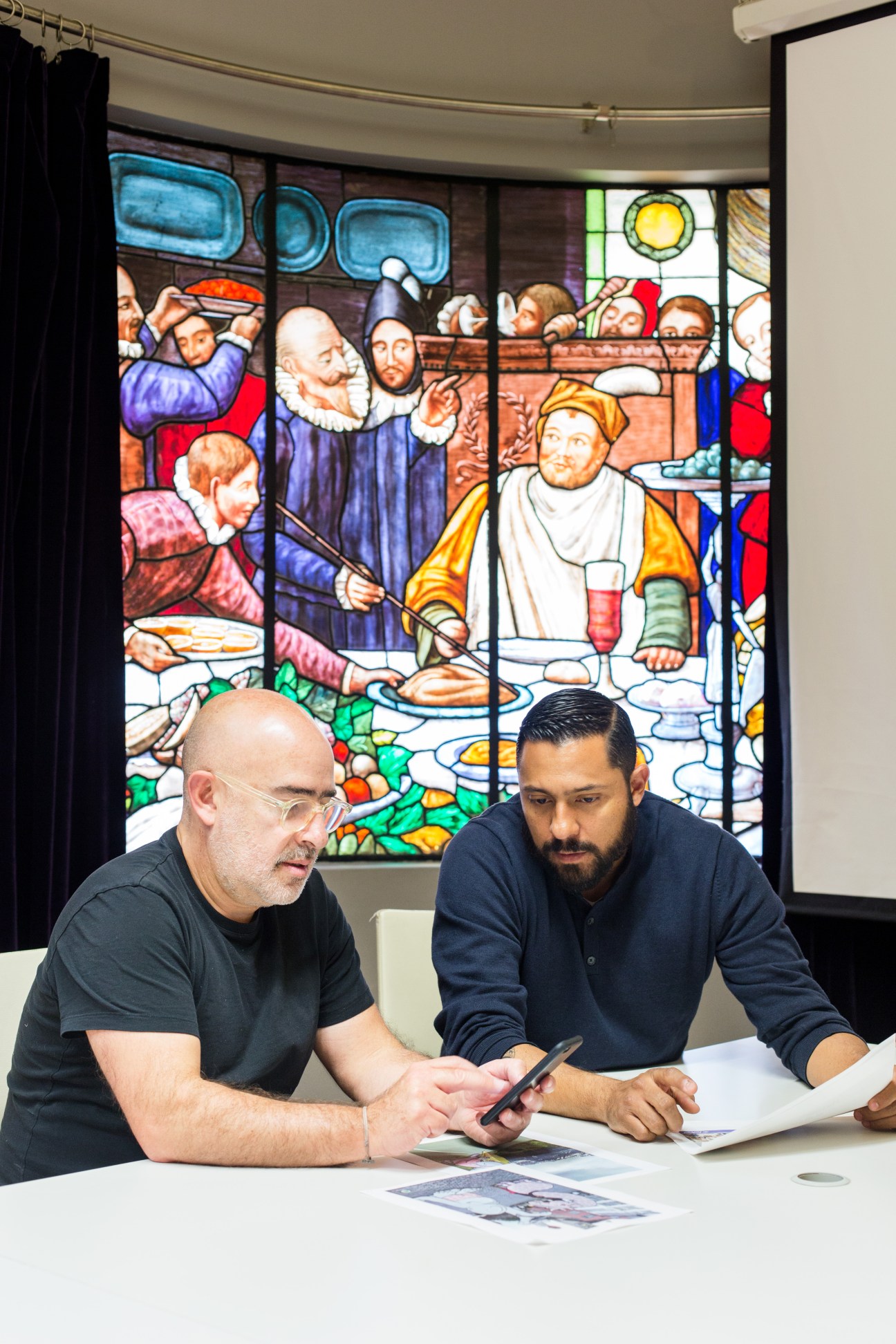 Raúl Cardós and Rafa Martínez review work at a table in front of stained glass religious artwork.