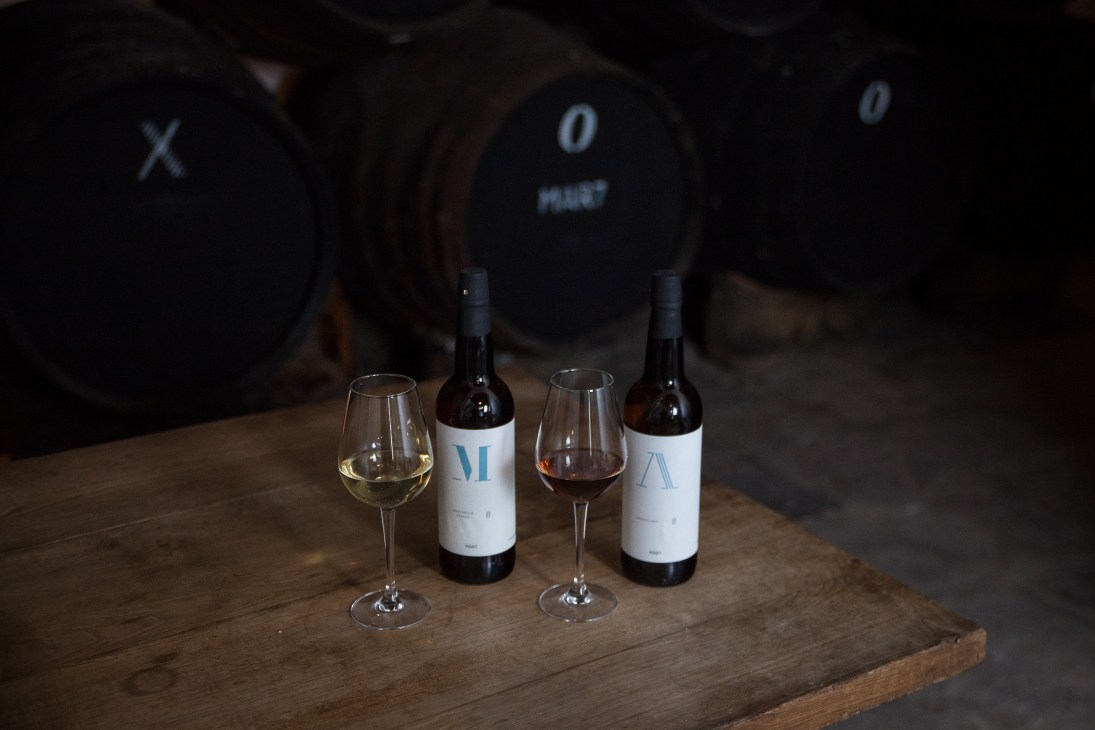 Two sherry bottles and filled wine glasses on a wooden table with aged barrels in the background.