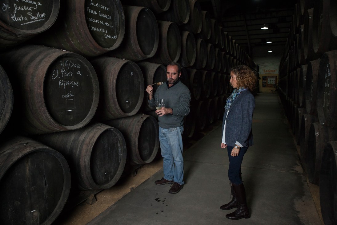 Two people stand in a cellar lined with large wooden barrels labeled with chalk markings at Despacho de Vinos Mar 7, Cádiz.