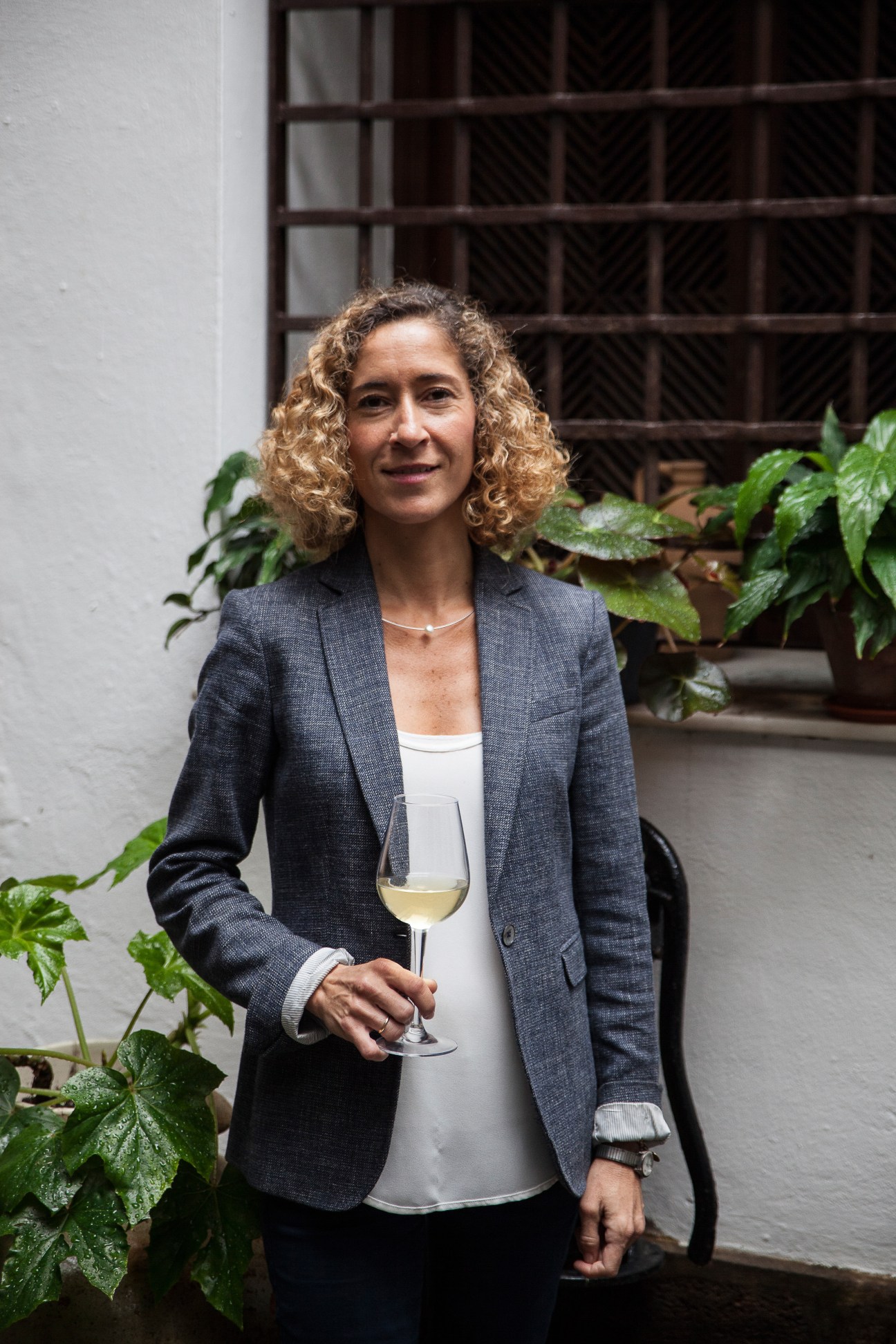 Woman with curly hair holding a glass of pale sherry, standing in a courtyard with green plants and a dark wooden latticed window behind her.