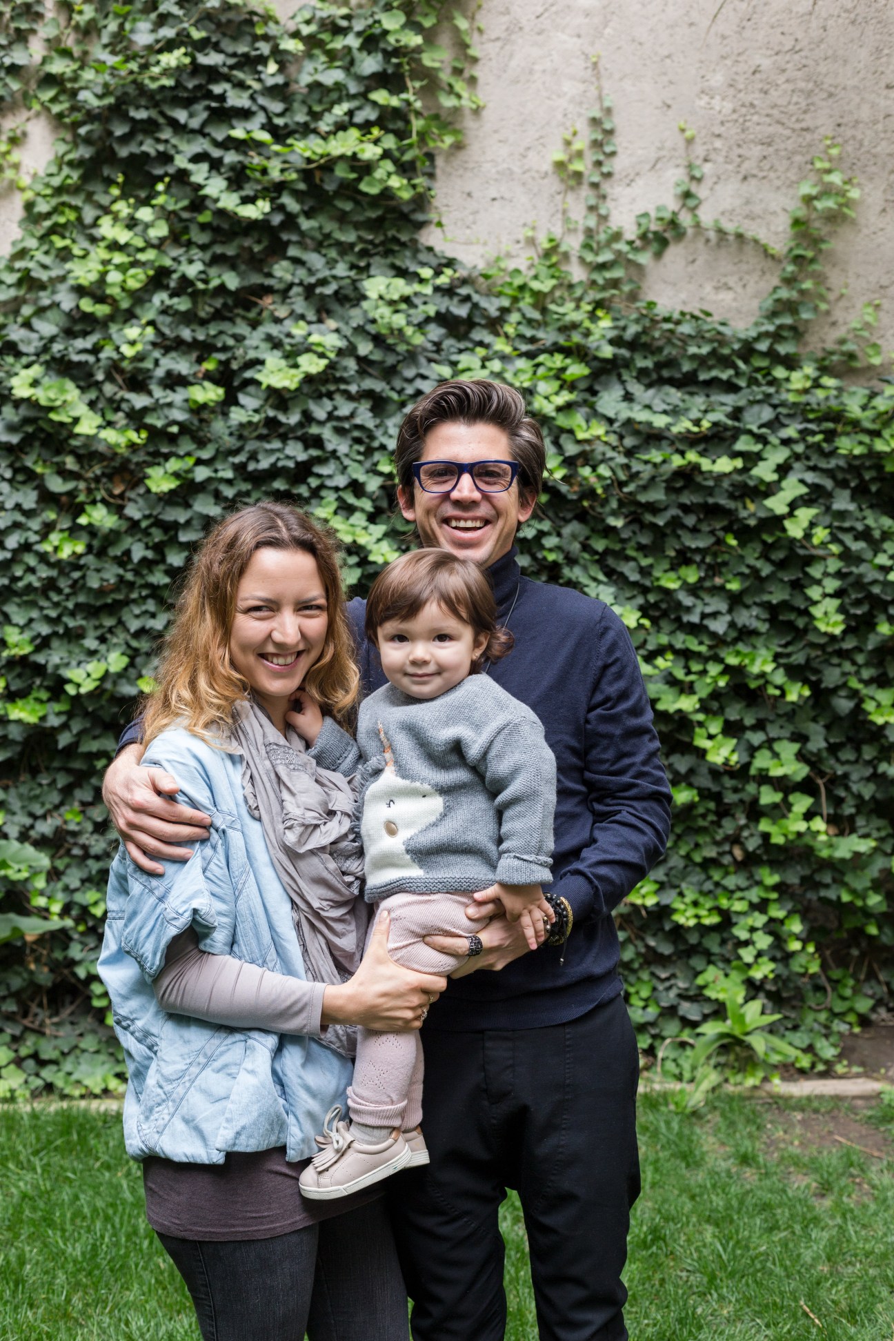Julio Amezcua stands with his wife and daughter in front of ivy-covered wall in a garden setting.