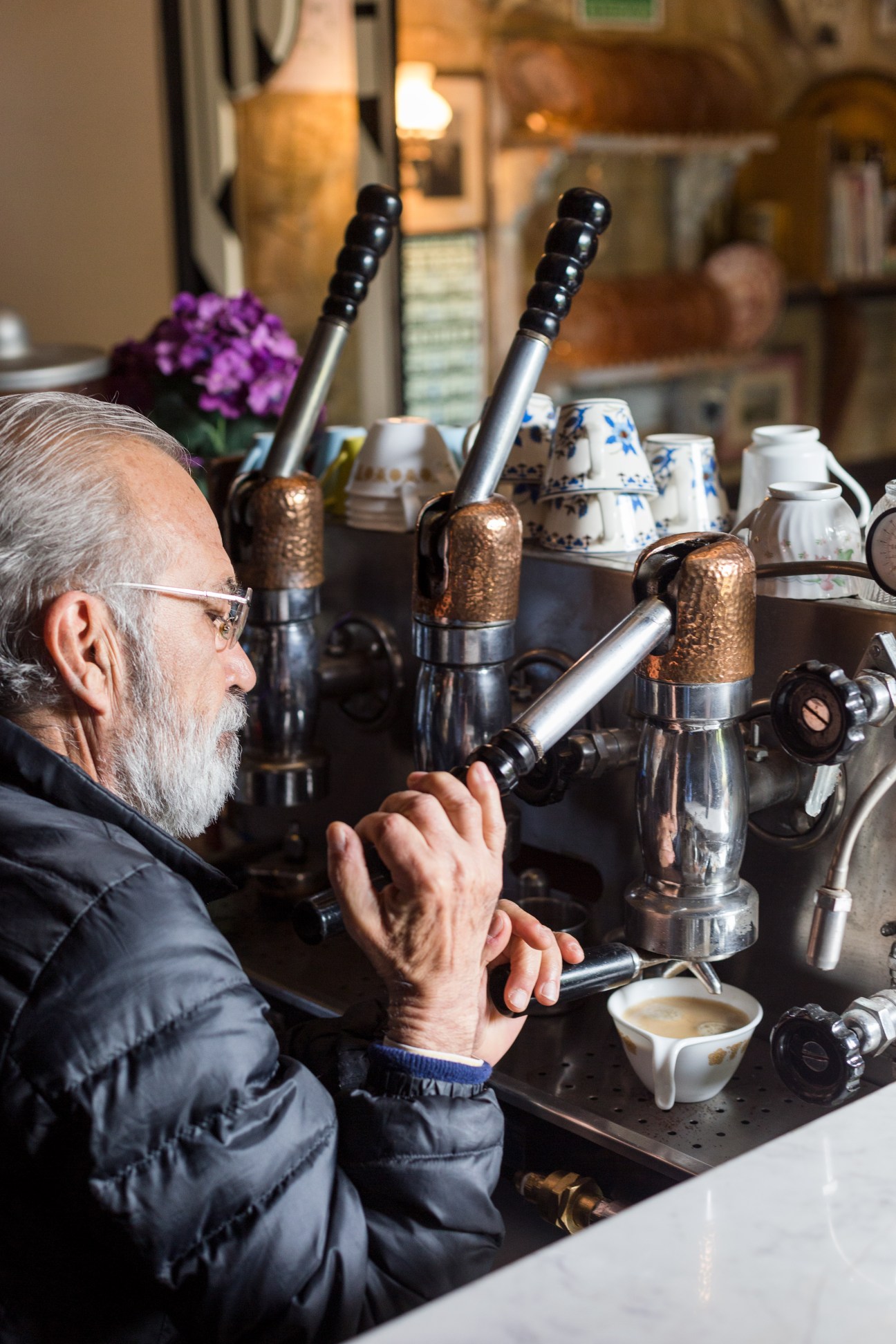 Older man with glasses pouring a drink at a bar with ornate brass taps and white porcelain cups.