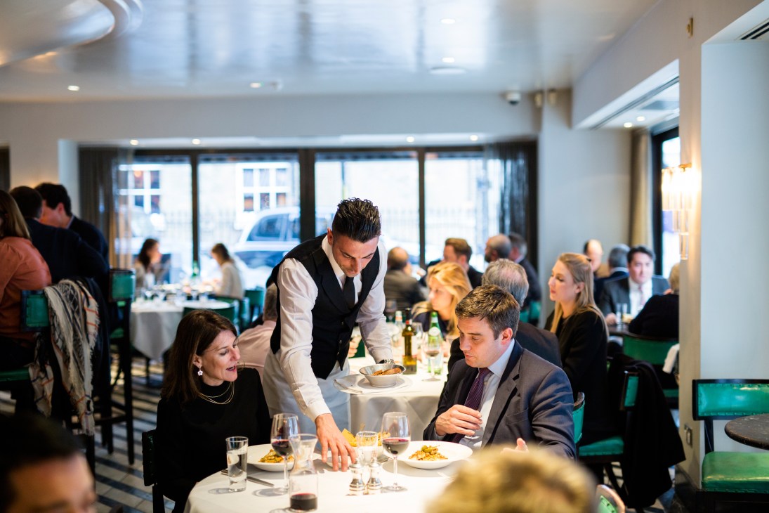 Waiter serving diners at a restaurant table in an elegant dining room with green banquettes.