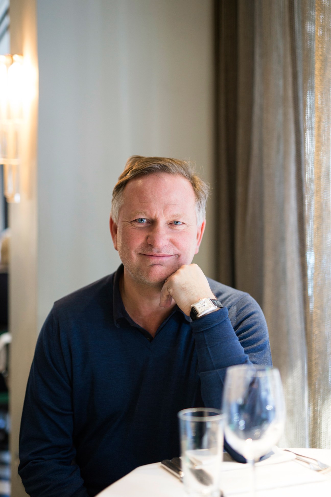 Nick Jones seated at a restaurant table, resting his chin on his hand, wearing a dark blue shirt.