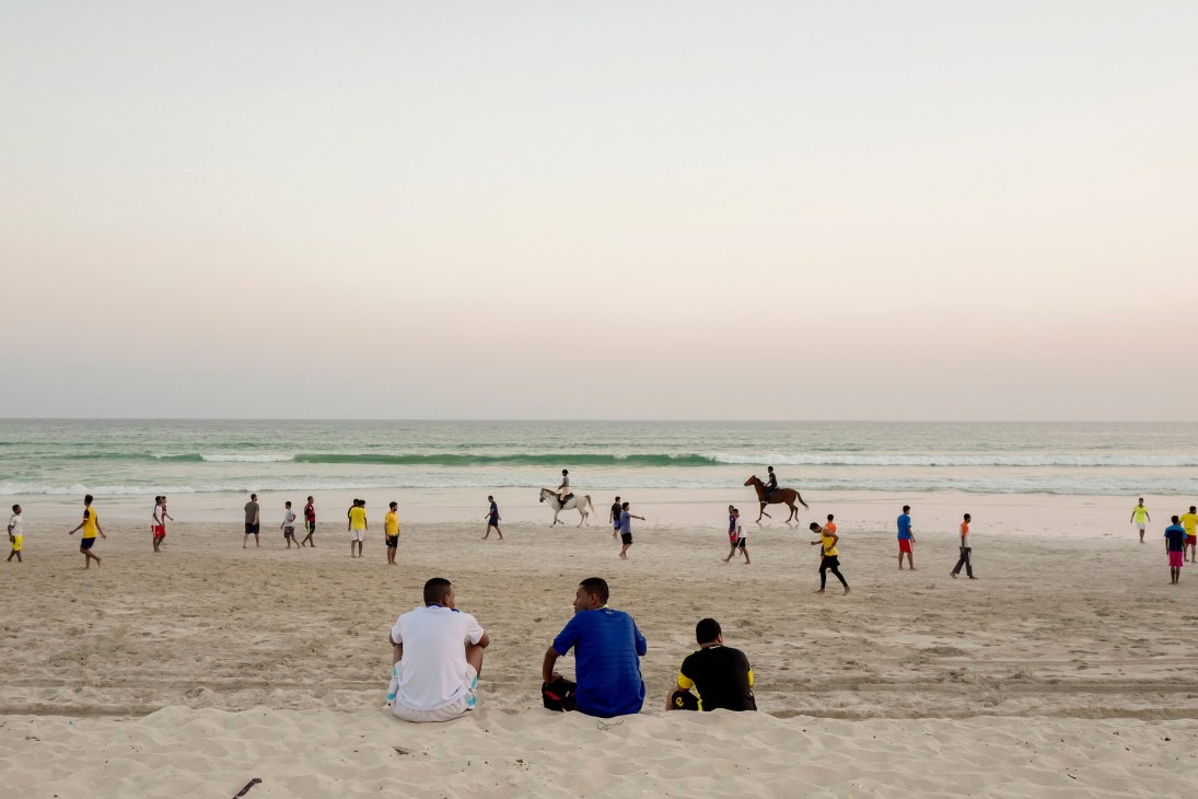 People enjoying a white sandy beach in Salalah with gentle waves and a horse rider visible in the water.
