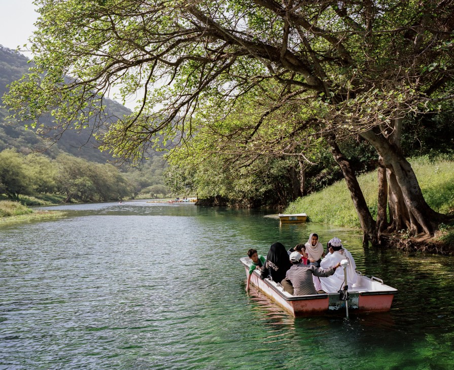 People in a boat on a lush green river lined with large trees and mountains in Salalah, Oman.