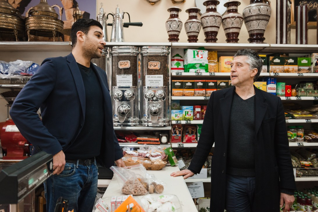 Two men conversing inside Harb Lebanese shop with shelves of packaged goods and decorative urns behind them.