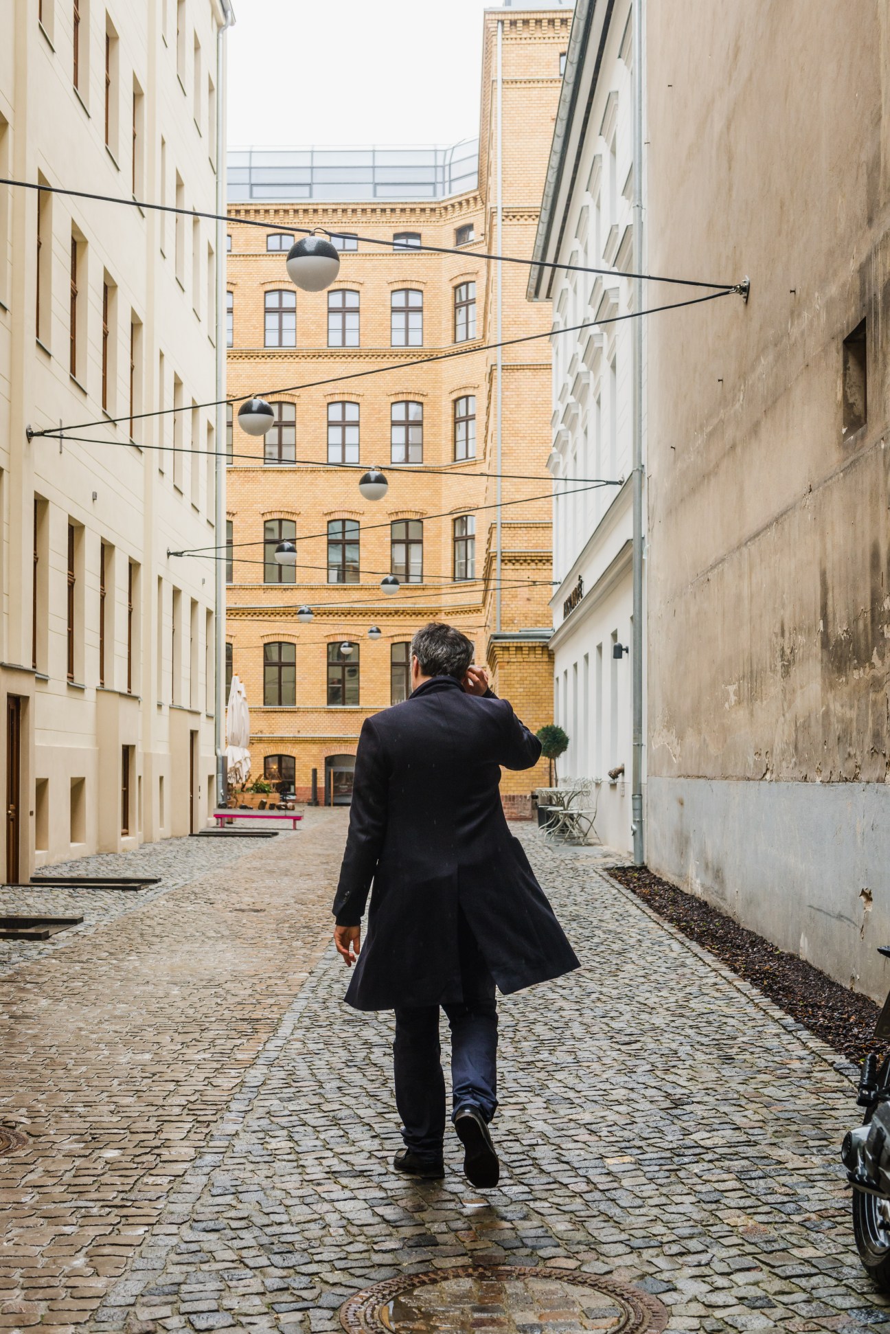 Man in black coat walking away through a cobblestone courtyard lined with brick buildings in Berlin.