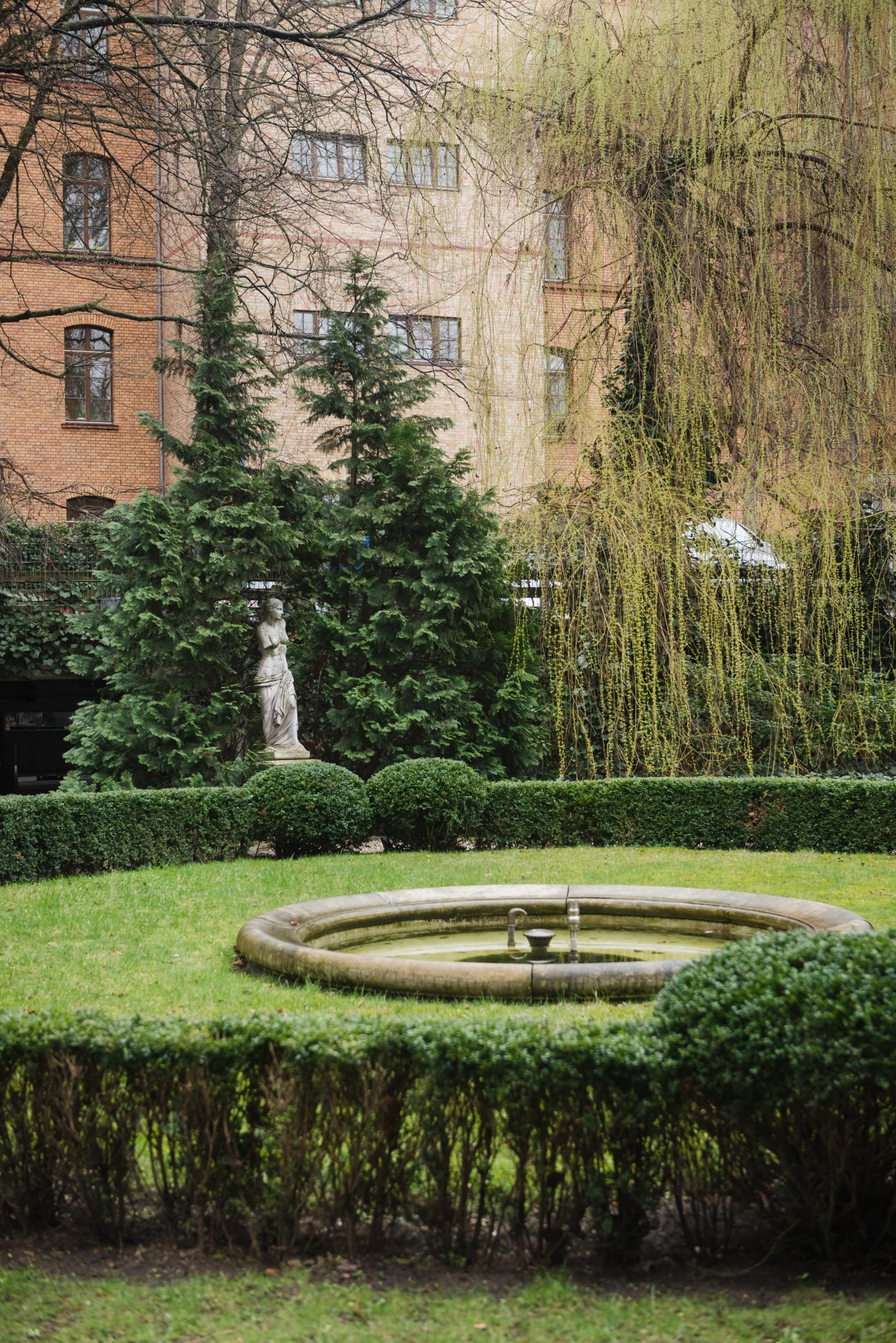 A manicured courtyard with a circular fountain, surrounded by hedges, with brick building and weeping willow tree in background.