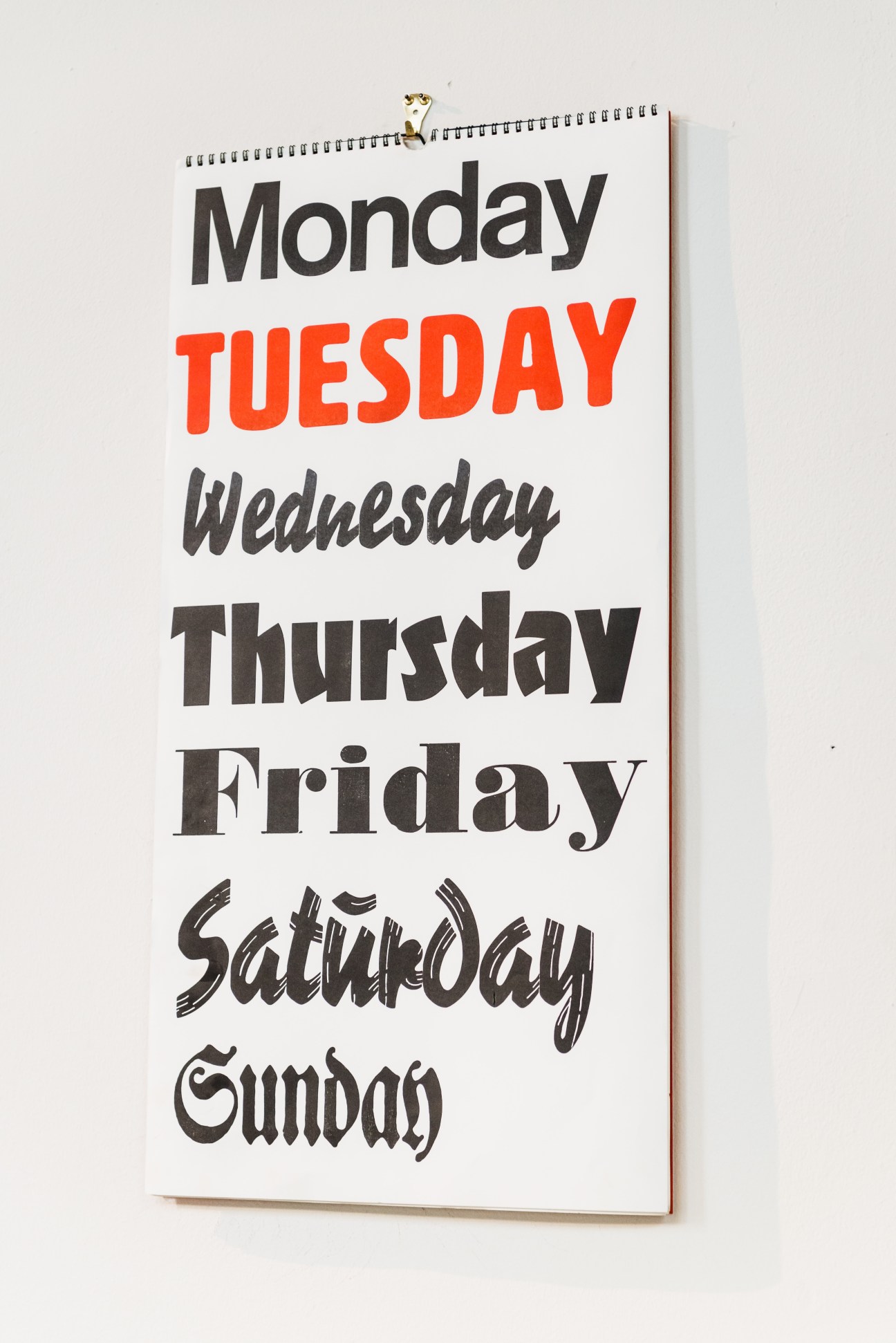 Wall-mounted calendar displaying days of the week in various fonts and colors, with Monday and Tuesday in black, Tuesday in bright red, and remaining days in dark text.