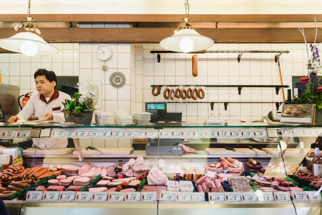 A butcher shop counter displays various cuts of meat and sausages behind glass with a staff member in a pink shirt standing behind the counter.