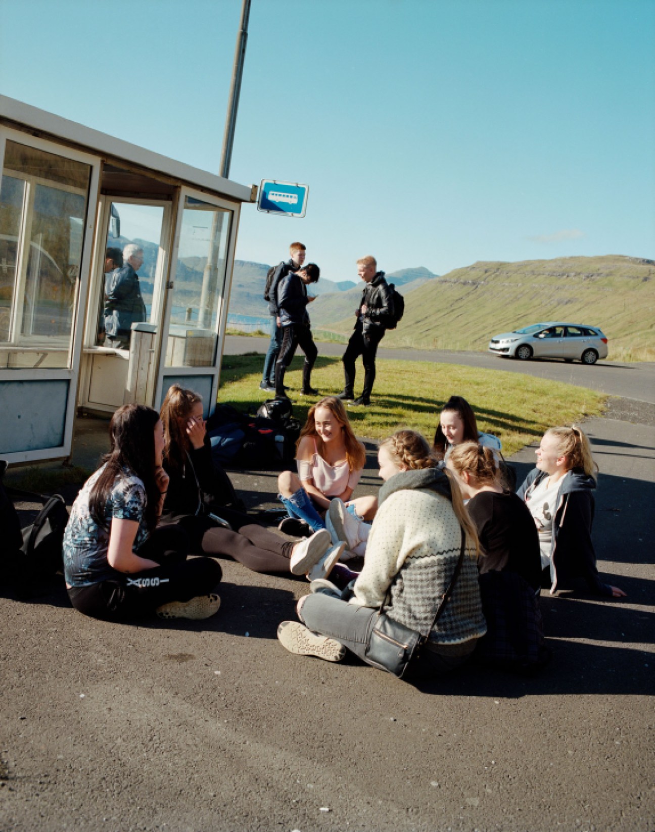 A group of people sit on a road beside a bus shelter with green mountains visible in the Faroe Islands landscape.
