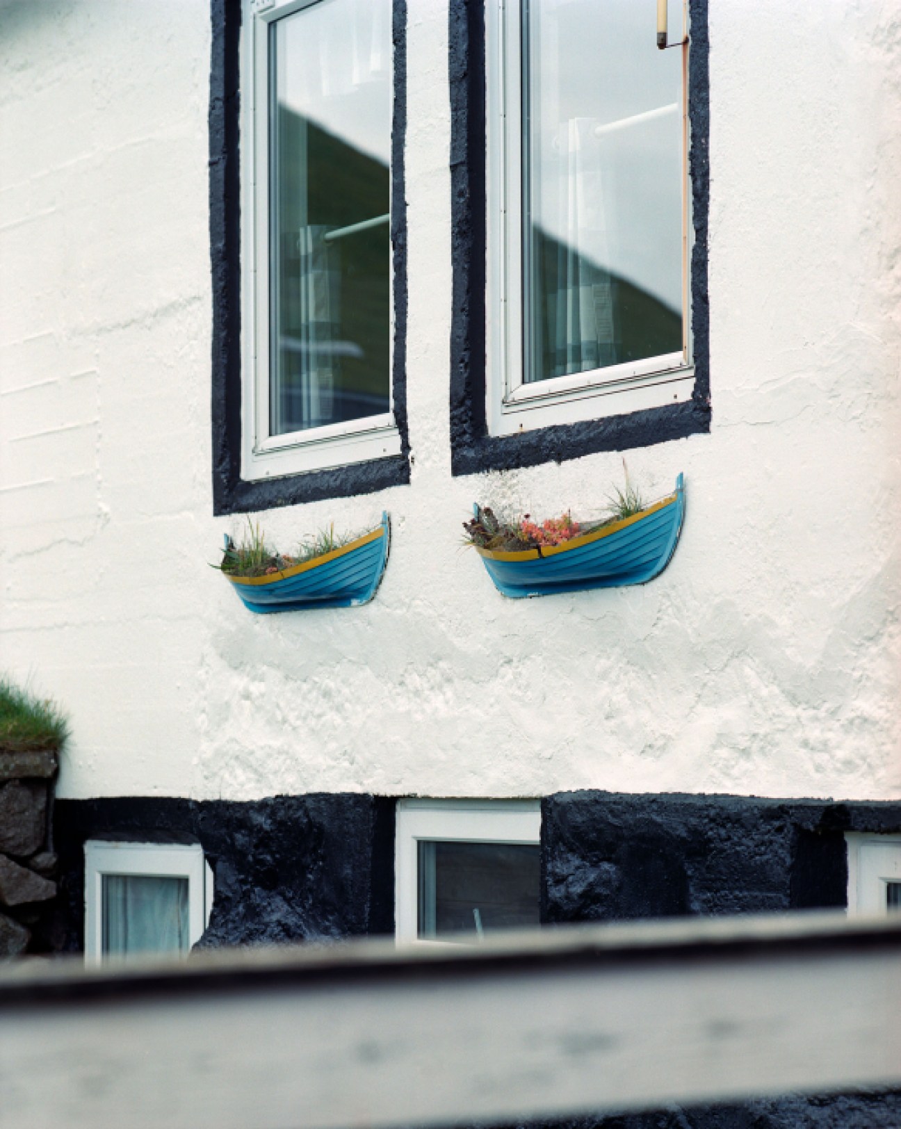 White-painted Faroese house with black trim, two windows with dark frames, and blue decorative boat planters beneath each window.