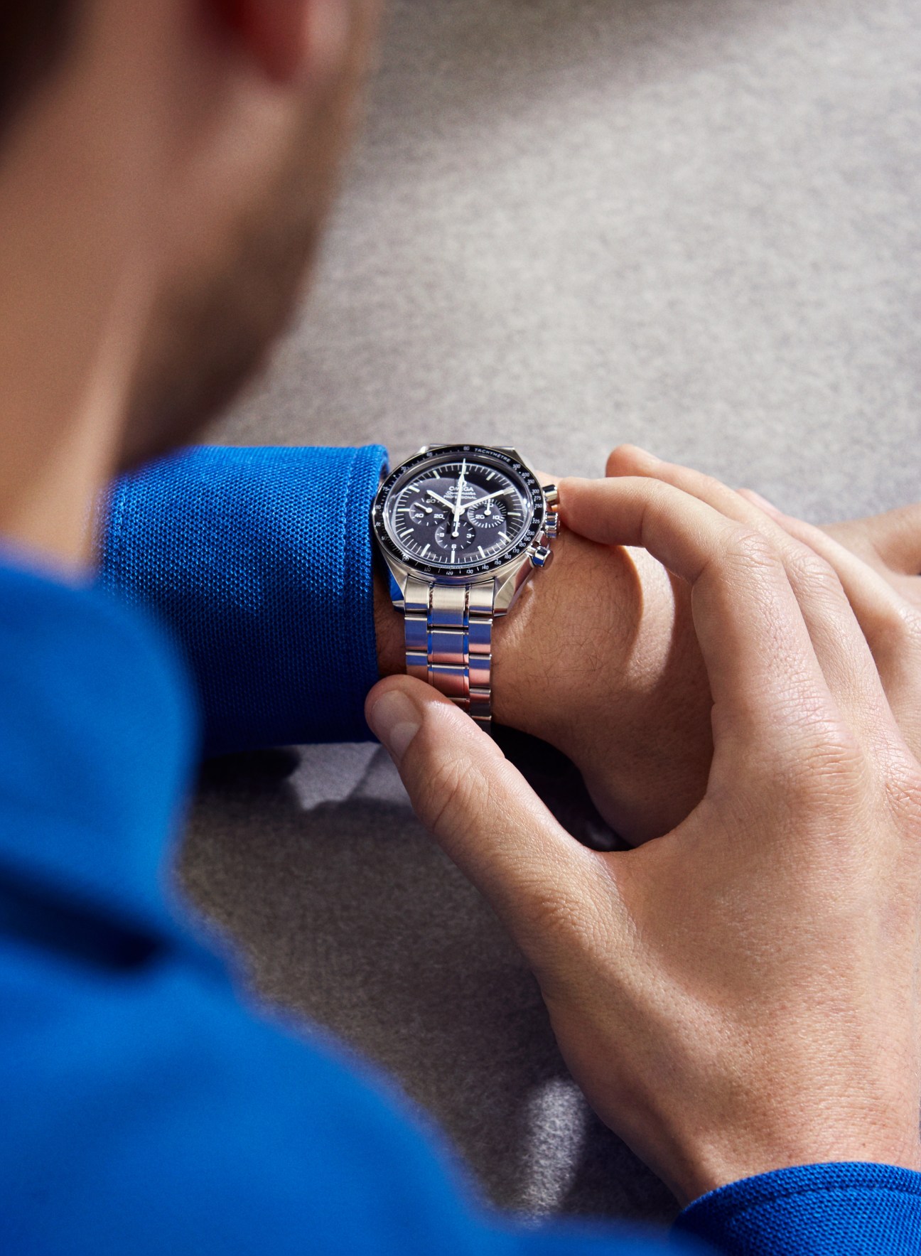 Person wearing Omega Speedmaster Moonwatch Professional chronograph with black dial and metal bracelet, blue sleeve visible.
