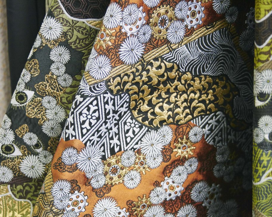 Close-up detail of an ornate fabric design featuring white daisy flowers, gold leaf patterns, and decorative geometric elements on dark and earth-toned backgrounds.