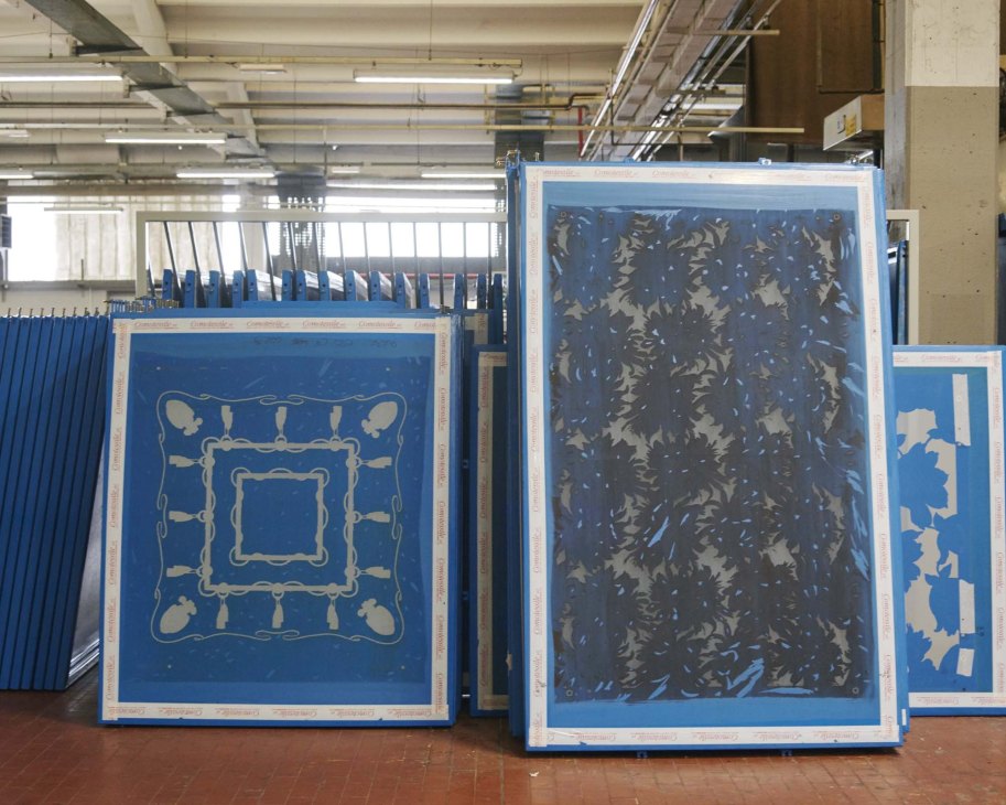 Large blue silk-screen printing frames with cream-colored designs displayed in an industrial factory workspace.