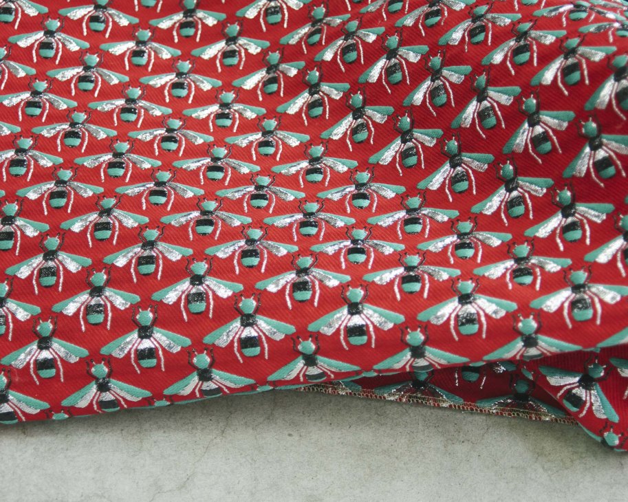 Close-up detail of a red silk fabric with repeating pattern of green and white stylized bees.