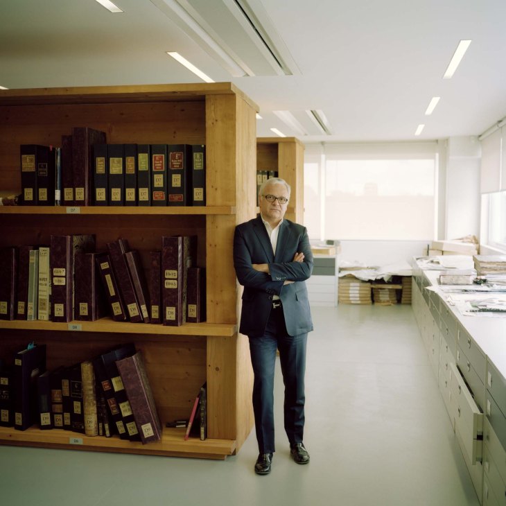 Francesco Gentili stands with crossed arms next to wooden shelving filled with catalogued fabric archives.