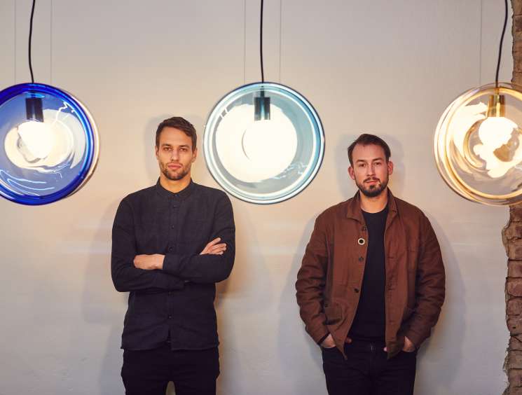 Two men stand against a white wall beneath three colorful pendant lights in blue, light blue, and amber tones.