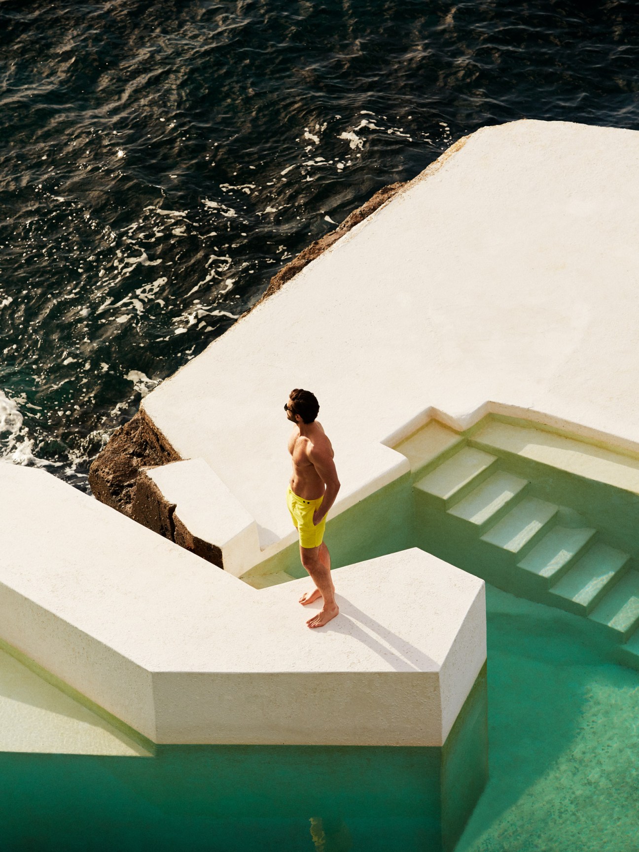 Man in yellow Frescobol Carioca swim shorts standing on white geometric architecture overlooking dark ocean water.