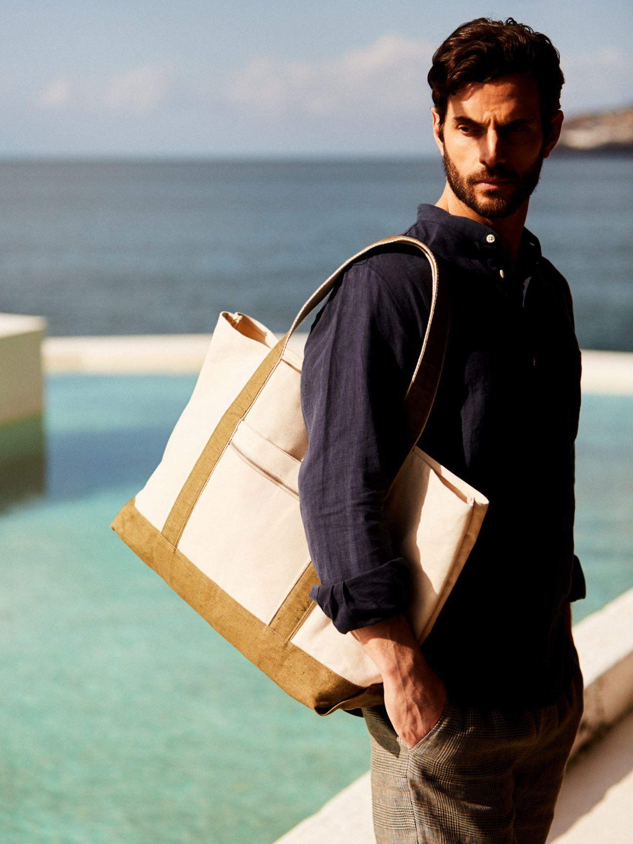 Man wearing a dark navy linen shirt and tan shorts holds a cream and natural jute tote bag by the seaside.
