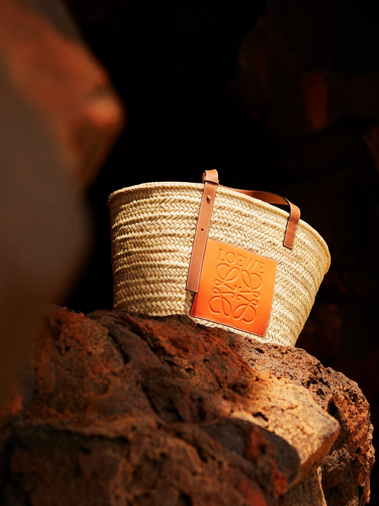 Loewe Paula's Ibiza woven straw basket bag with orange leather handle and brand tag on rocky surface.