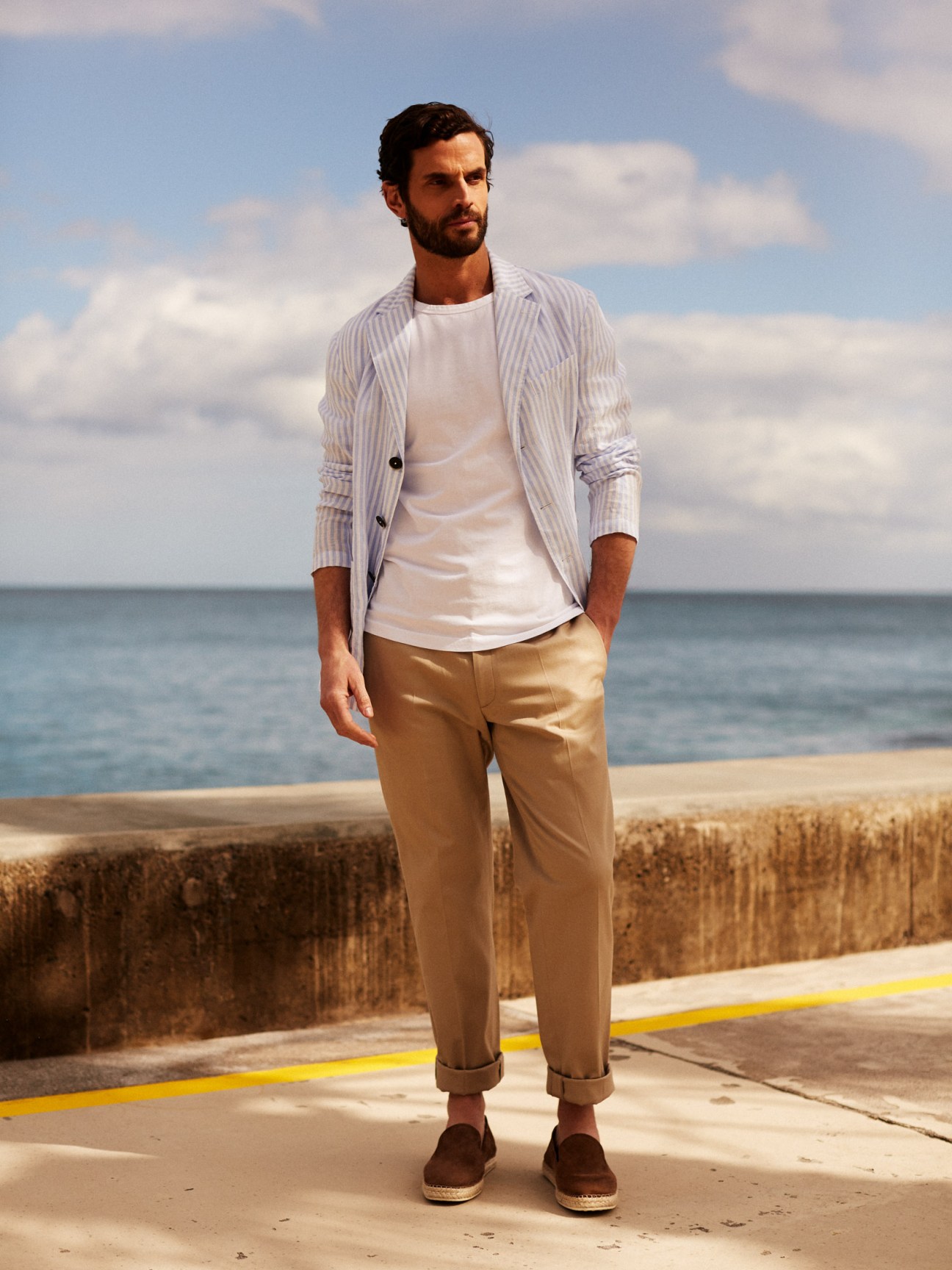 Man wearing a striped shirt, white t-shirt, tan trousers and brown slip-ons standing on a beach promenade.