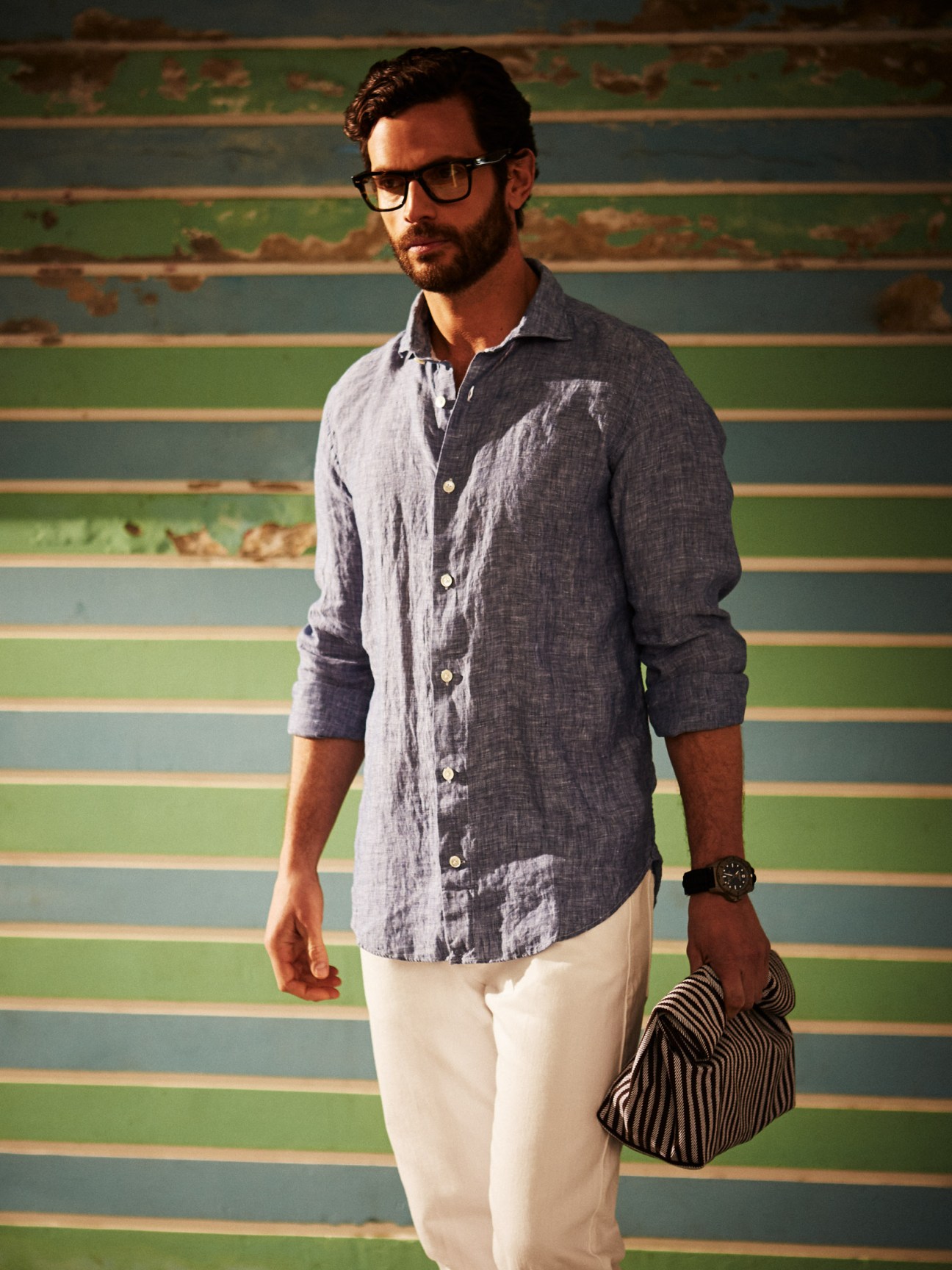 Man wearing Eleventy shirt and jeans, Oliver Peoples glasses, Victorinox watch, and Steve Mono bag against striped background.