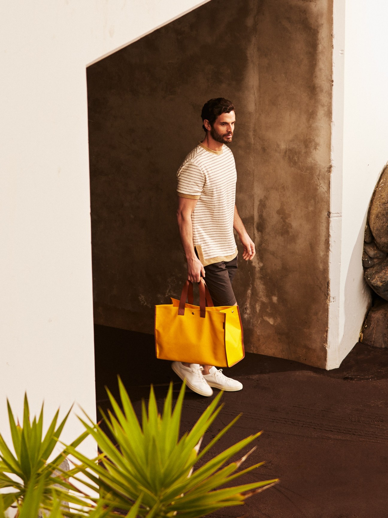 Man wearing striped t-shirt by The Gigi, shorts by Closed, CQP trainers, and yellow L/Uniform bag.