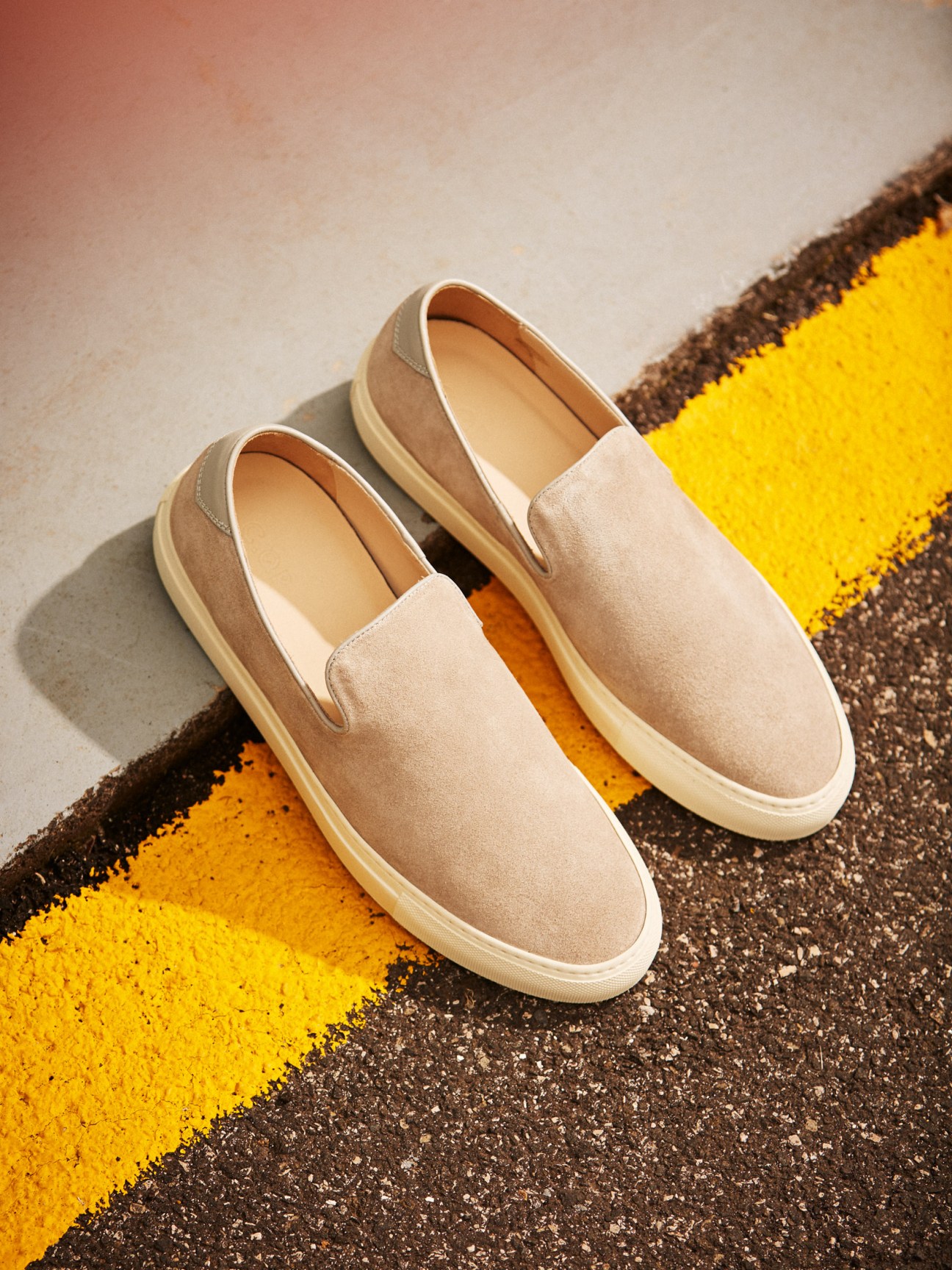 Beige suede slip-on sneakers with cream rubber sole on yellow and black striped pavement.
