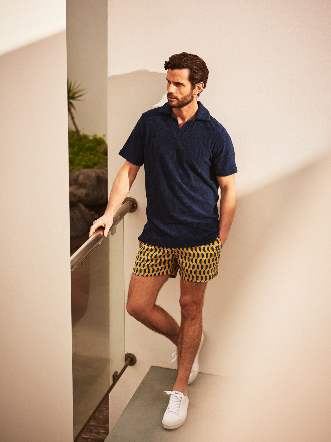 Man wearing navy polo shirt by The Gigi, yellow patterned swim shorts by Timo, and white Spring Court trainers on a minimalist staircase.
