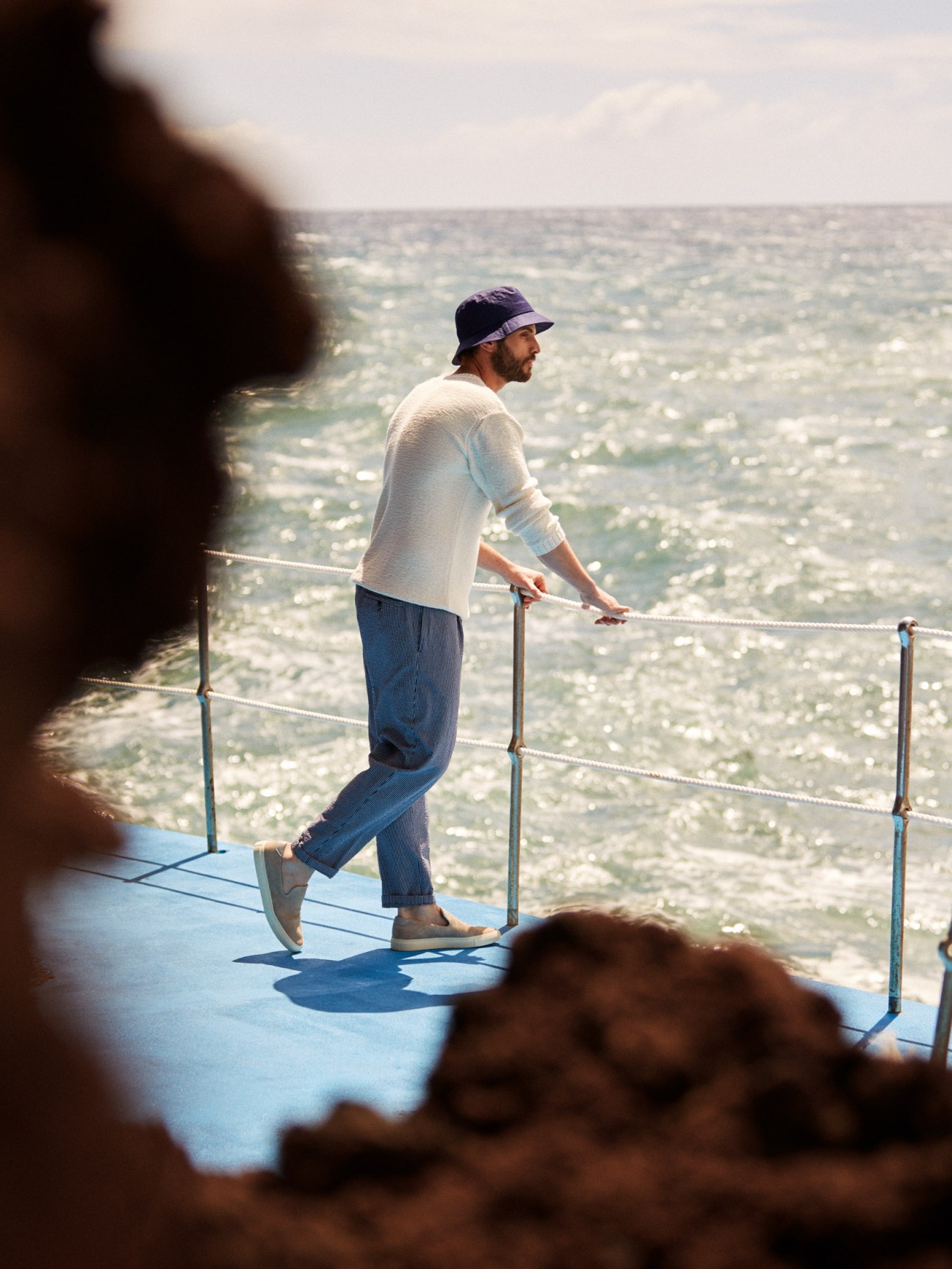 Man in cream jumper by Closed, navy trousers by The Gigi, and La Paz hat standing by seaside railing overlooking ocean.
