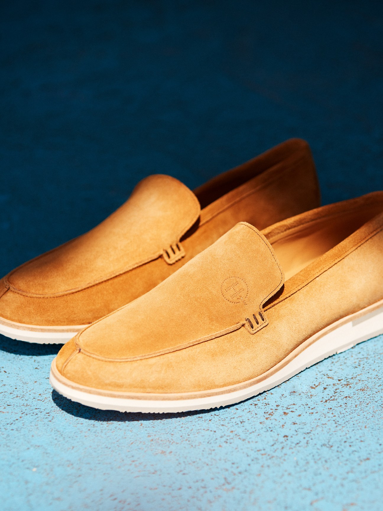 Three tan suede loafers with white soles displayed on a blue textured surface against a deep blue background.