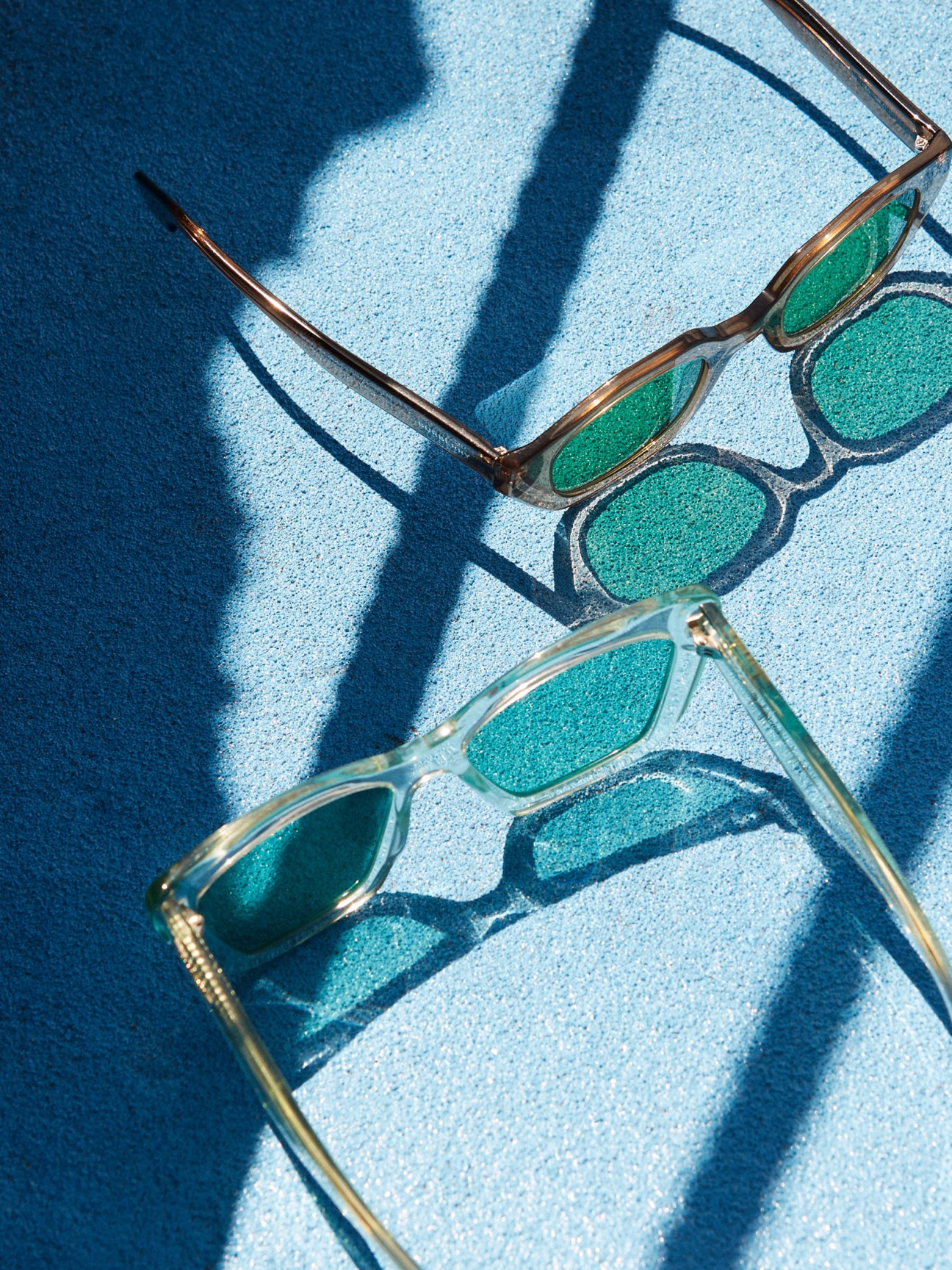 Three pairs of sunglasses with turquoise and green lenses arranged on a light blue textured surface casting shadows.