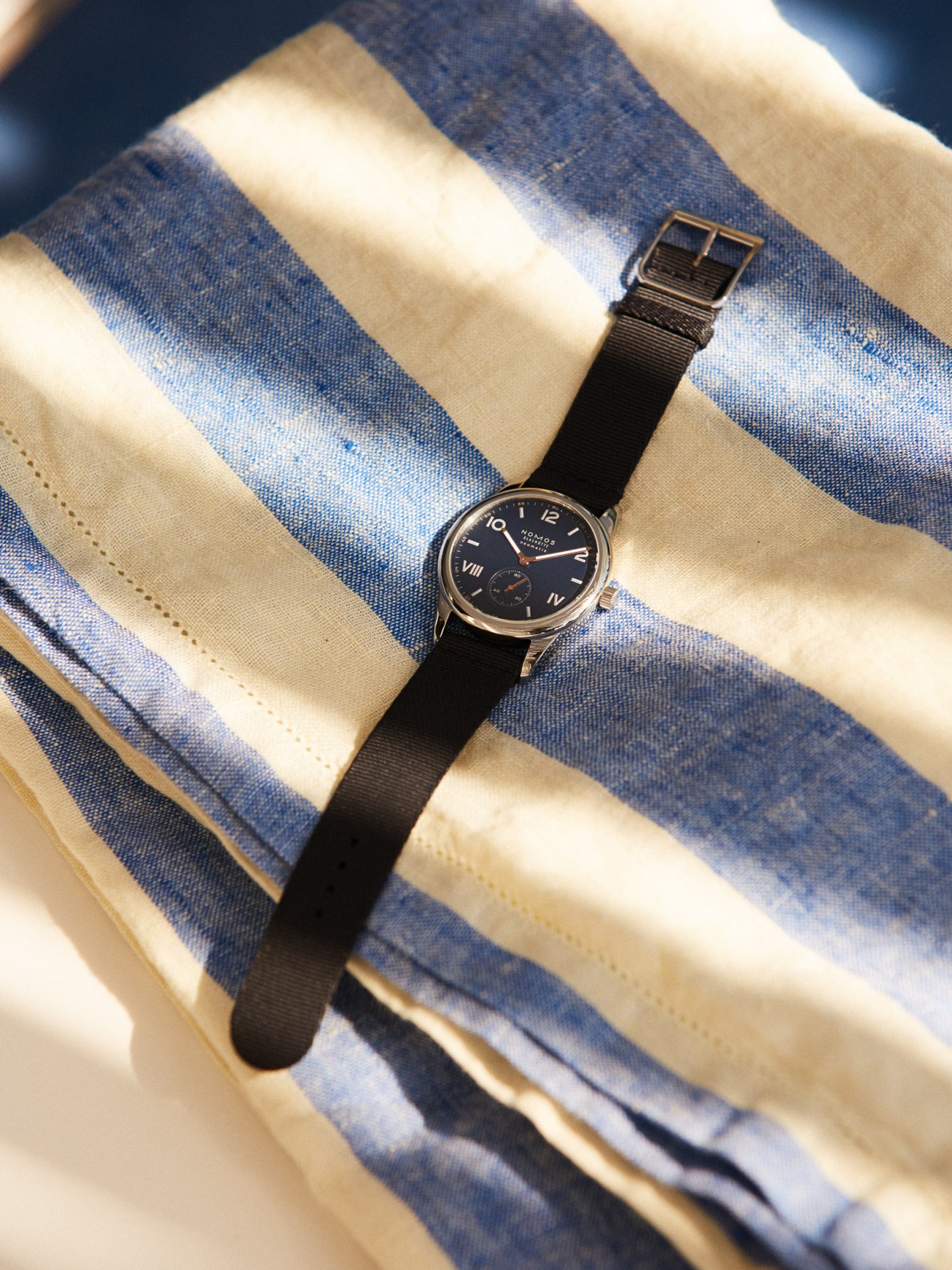 Nomos Glashütte watch with black leather strap on blue and white striped Frescobol Carioca beach towel.