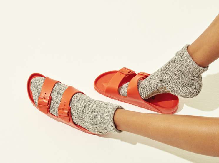 Red Birkenstock sandals worn with grey knit socks on two legs against a cream background.