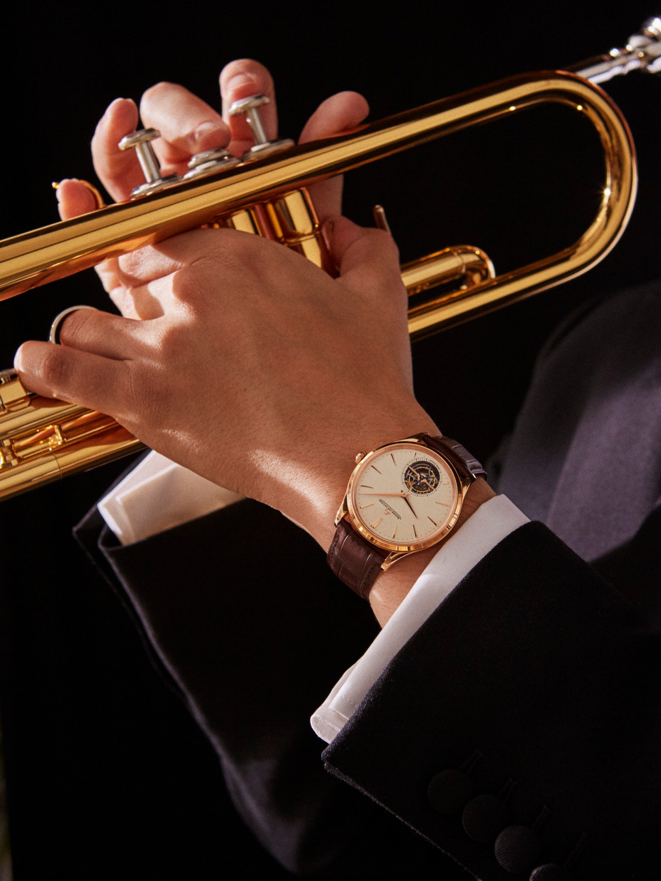 A hand holding a brass trumpet wearing a Master Ultra Thin Tourbillon watch by Jaeger-LeCoultre with a brown leather strap.