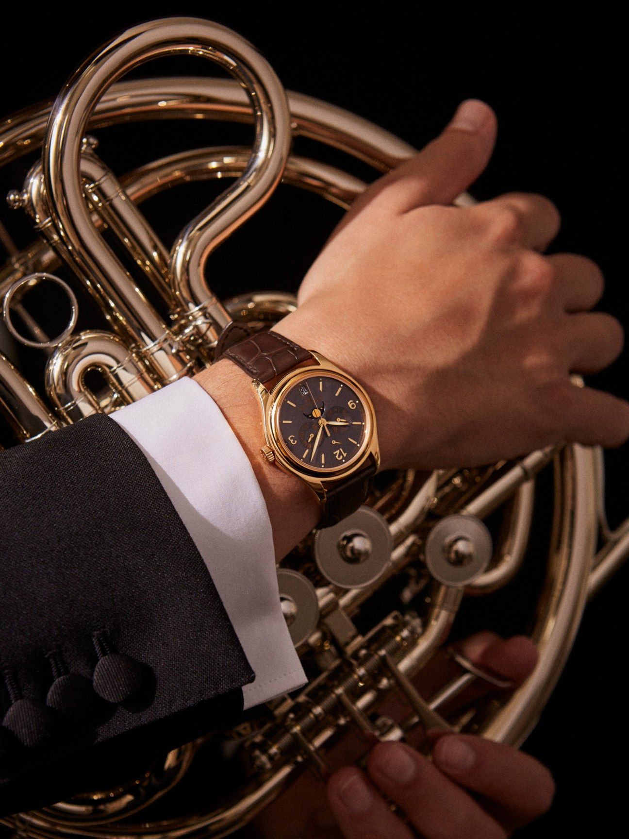 Wrist wearing Patek Philippe 5146j watch with brown leather strap, holding brass instrument against black background.