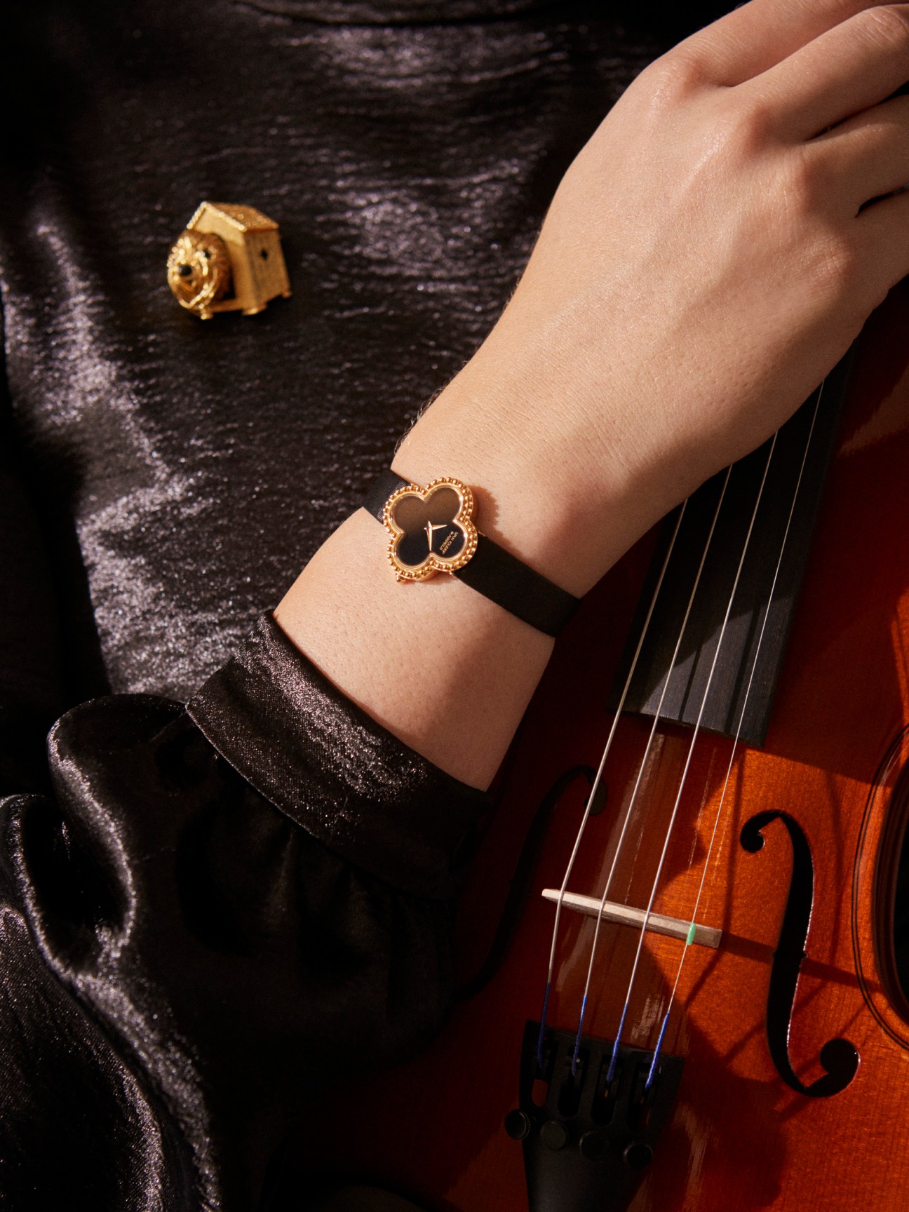 Van Cleef & Arpels Alhambra watch worn on wrist with matching brooch, styled with violin and sparkly fabric.
