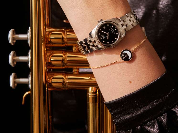 Tudor Glamour Date 26 watch with steel bracelet worn on wrist next to gold trumpet