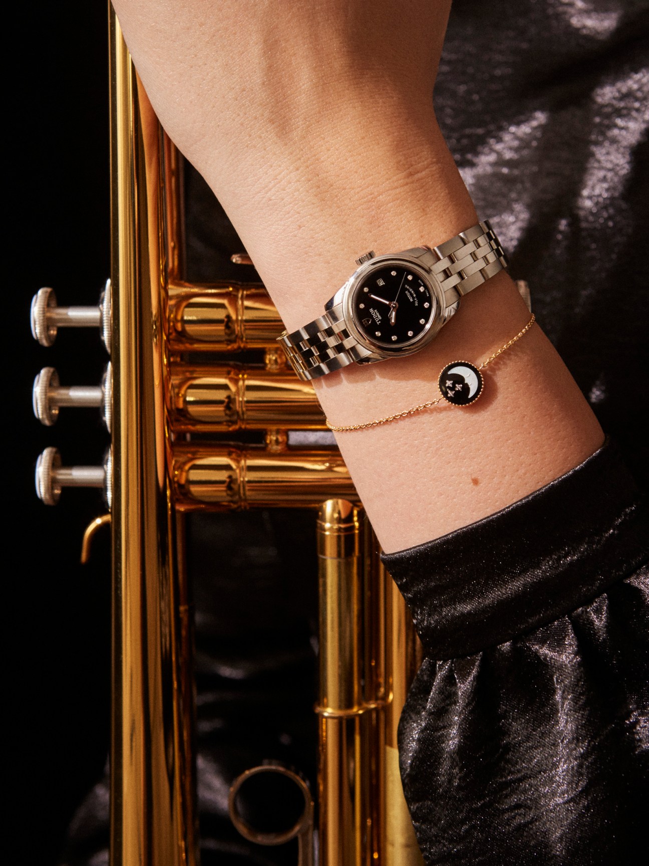 Tudor Glamour Date 26 watch with steel bracelet worn on wrist next to gold trumpet