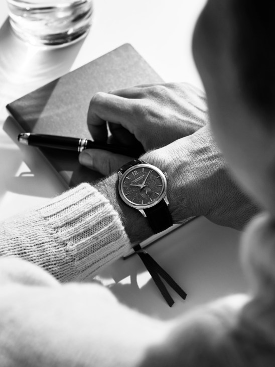 Black and white photograph of a wrist wearing a Chopard LUC XPS watch and cream sweater, holding a Montblanc fountain pen over a diary.