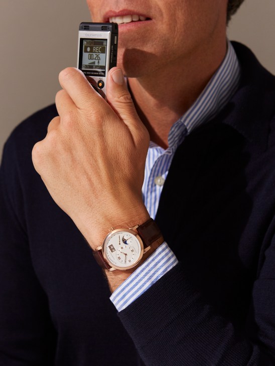 Man in striped shirt and cardigan holding Olympus recording device, wearing A Lange & Söhne moon phase watch.
