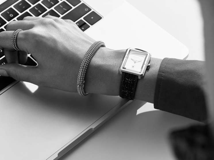 Close-up black and white image of a woman's wrist wearing a Chanel Watches Boyfriend watch, Van Cleef & Arpels bangles and ring, typing on a laptop.