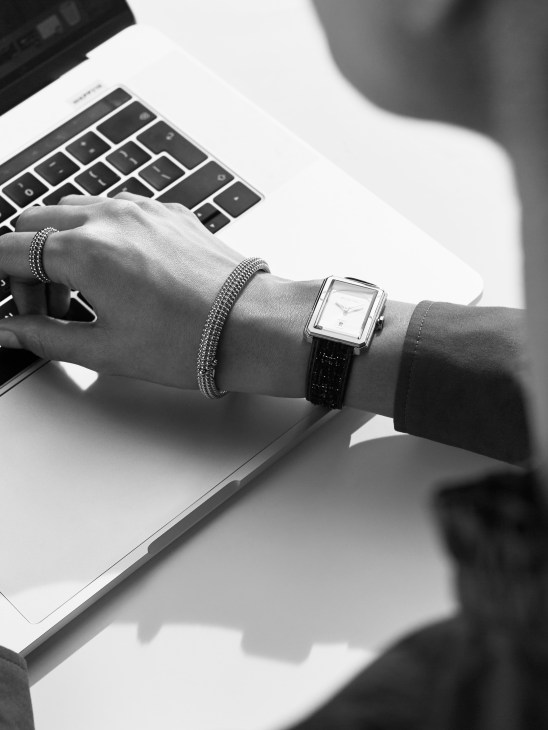 Close-up black and white image of a woman's wrist wearing a Chanel Watches Boyfriend watch, Van Cleef & Arpels bangles and ring, typing on a laptop.