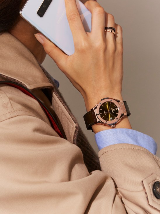 Hand holding a white Samsung smartphone wearing a Hublot Classic Fusion King Old Green watch and Chopard rings.
