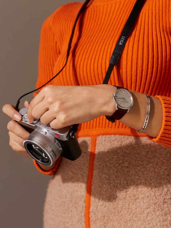 Woman in orange dress holding Leica camera, wearing Vacheron Constantin watch and Bulgari bangle.
