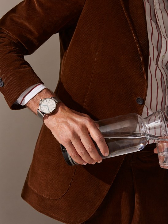 Man in brown corduroy suit holding a glass, wearing a Nomos Glashütte watch and striped shirt.