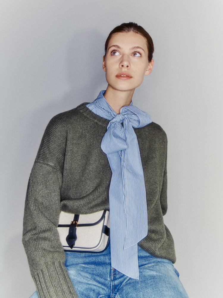 Woman in gray knit sweater and blue striped bow-tie shirt looking upward, wearing jeans and white belt bag