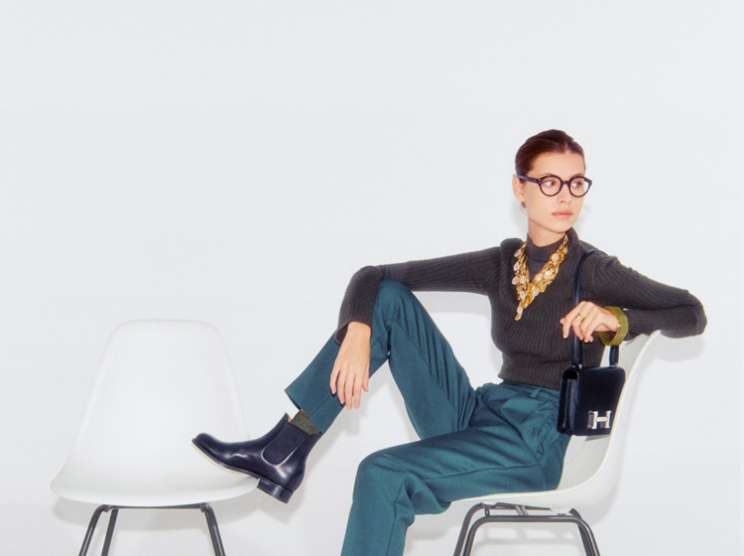 Woman in teal trousers and dark sweater sits on white chair, wearing round glasses and holding small black handbag